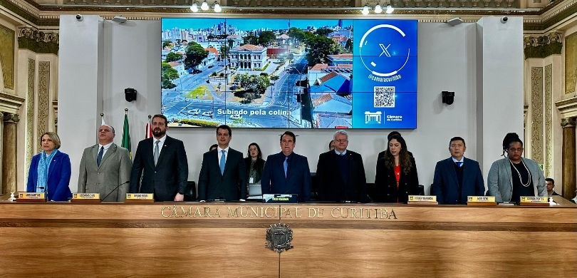 Fotografia em que se vê a mesa do plenário da Câmara Municipal de Curitiba, na qual aparecem em ...