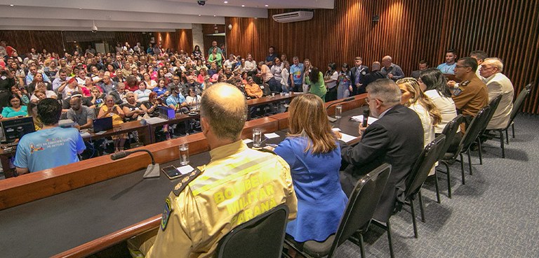 A foto mostra a mesa da audiência pública, são oito pessoas sentadas, homens e mulheres, com mic...