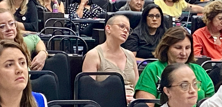 Fotografia de várias mulheres sentadas em cadeiras pretas, em um auditório. Em destaque, é possí...