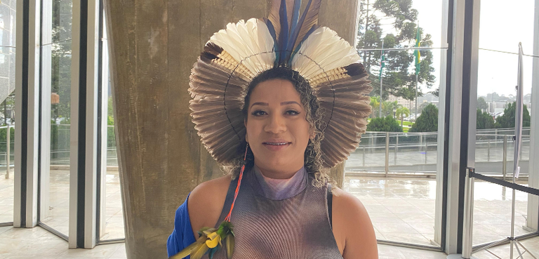 Fotografia de uma mulher indígena sorrindo. Ela tem cabelos cacheados, veste uma camisa de cores...