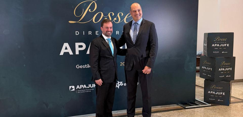 Fotografia de dois homens adultos, de pele clara, vestindo ternos escuros, camisas claras e grav...