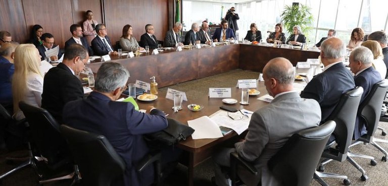 Fotografia de diversas pessoas, entre homens e mulheres, sentadas ao redor de uma grande mesa de...