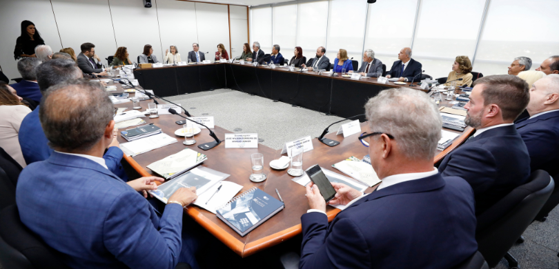 Fotografia de uma sala de reuniões institucional onde diversas pessoas, homens e mulheres de ida...
