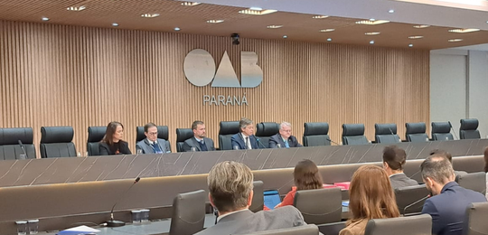 Fotografia que retrata um plenário institucional durante um evento. Ao fundo, centralizado em um...