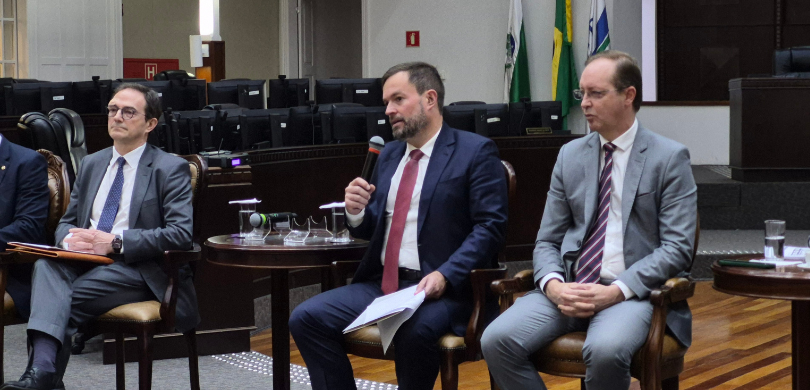 Fotografia em que se observam três homens sentados lado a lado em cadeiras de madeira com estofa...