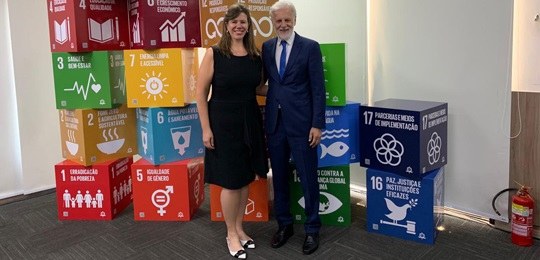 Fotografia de duas pessoas em pé: uma mulher (à esquerda) e um homem (à direita). Atrás deles, h...