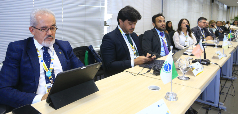 Fotografia em que se observa um grupo de pessoas, entre homens e mulheres, sentados ao longo de ...