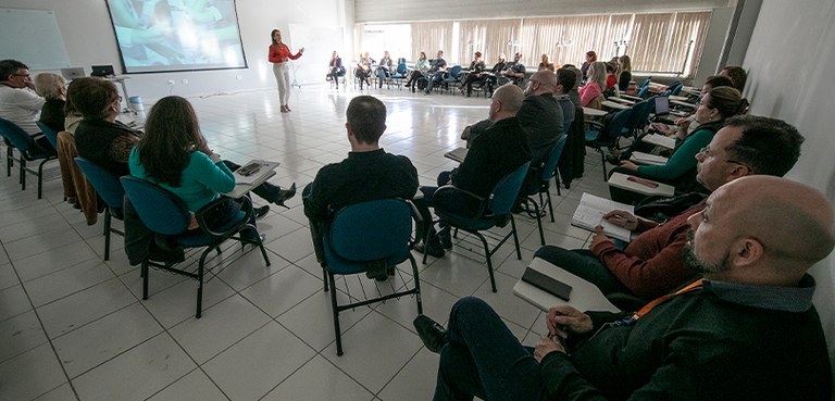 Fotografia de uma sala com diversas pessoas sentadas em carteiras escolares, organizadas em meio...