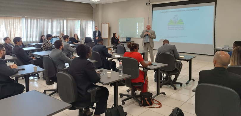 Fotografia de um homem de terno segurando um microfone enquanto fala diante de uma plateia, form...