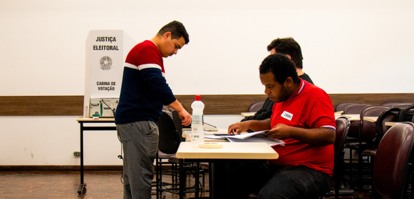 Fotografia em que se observam três homens em um ambiente interno organizado para votação. Em pri...
