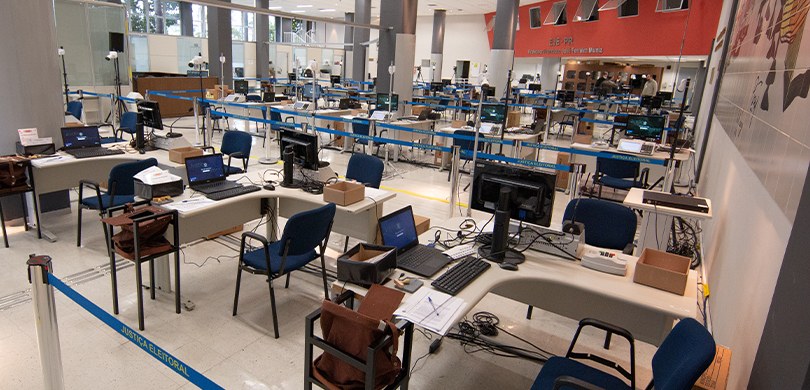 Fotografia de uma sala grande, com diversas mesas, urnas eletrônicas cadeiras e computadores.