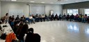 Fotografia de várias pessoas em uma sala, sentadas em cadeiras organizadas em círculo. Ao redor,...
