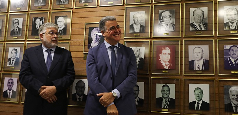 Fotografia de dois homens de terno posando sorrindo diante de uma parede marrom, com fotos em pr...