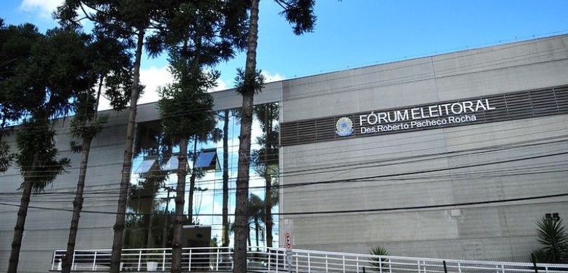 Na imagem, vê-se a fachada do Fórum Eleitoral de Curitiba. O edifício é cinza e possui uma estru...