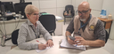 Fotografia em que se observam duas pessoas sentadas ao redor de uma mesa redonda clara, em um es...