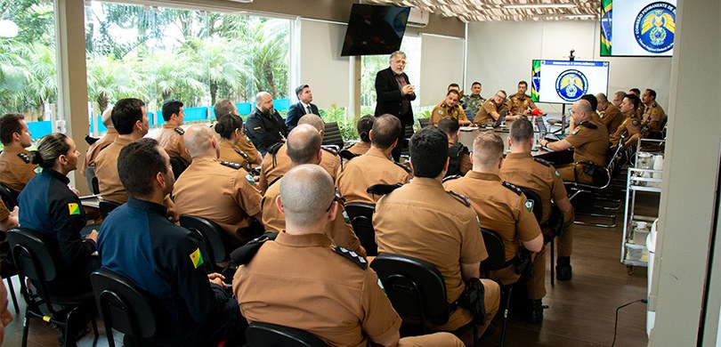 TRE-PR recebe oficiais da Polícia Militar para curso de formação Em uma sala envidraçada, diversos homens fardados estão sentados, ouvindo um homem, vestindo rou...