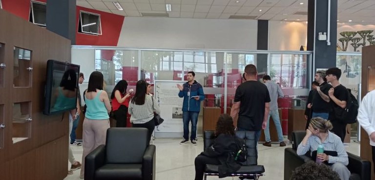 TRE-PR recebe visita de acadêmicos de Direito da FAPAR 2 Na imagem veem-se pessoas espalhadas em um saguão. Um grupo está de pé, ao redor de um homem que...