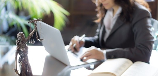 TRE-PR reduz prazos relativos às sessões virtuais de abril Fotografia de uma balança da Justiça, em primeiro plano, e de uma mulher em frente a um notebook...