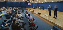 Fotografia que retrata um auditório amplo durante um evento institucional. No palco, à direita, ...