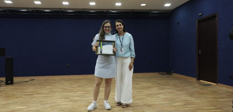 TRE-PR reinaugura auditório em solenidade com homenagens à embaixadora de acessibilidade Pietra Silvestri e ao Arquivo Público 3 Fotografia de duas mulheres de pele clara posando em pé ao centro de um auditório. A mulher à es...