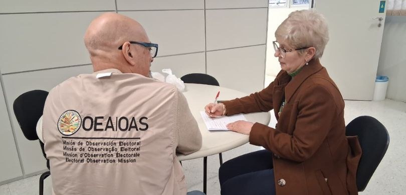 Em um ambiente interno de paredes claras, duas pessoas estão sentadas em uma mesa redonda branca...