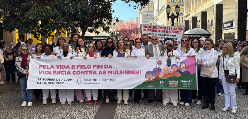 Fotografia de diversas pessoas, a maioria mulheres, segurando juntas uma faixa branca em que se ...