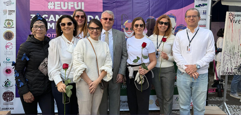 Fotografia de seis mulheres e dois homens posando sorrindo em frente a um banner roxo, em que é ...