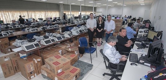 TRE-PR sedia teste em campo nacional dos sistemas eleitorais Fotografia em uma sala repleta de urnas eletrônicas dispostas em cima de bancadas sob as quais h...
