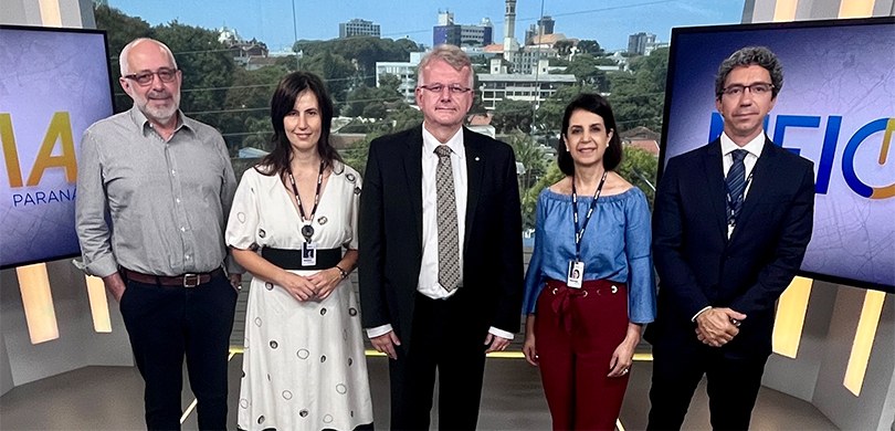 Fotografia de três homens e duas mulheres em um estúdio, em que se veem duas telas com o logo do...