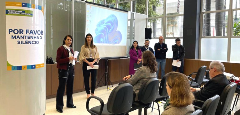 Foto em que aparecem seis pessoas em pé, do lado esquerdo, e três pessoas sentadas em cadeiras p...