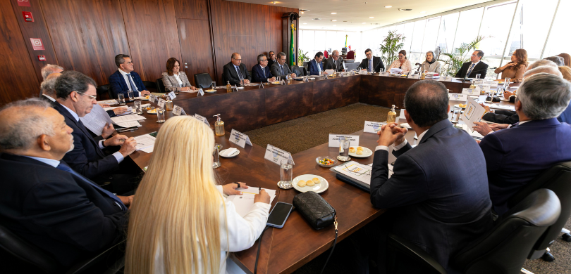 Fotografia de diversas autoridades, entre homens e mulheres, sentadas em uma mesa de reunião mar...