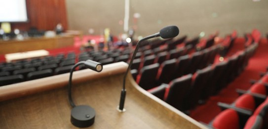 Imagem focada em um microfone de mesa preto e ao fundo um auditório com poltronas vermelhas