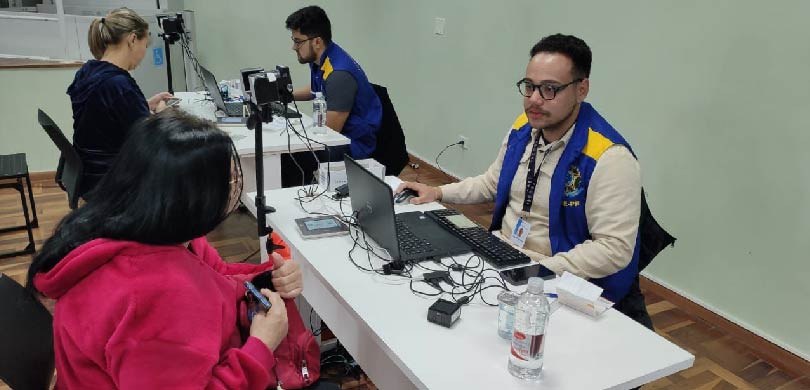 Fotografia de dois homens vestindo coletes azuis e amarelos da Justiça Eleitoral. Eles estão sen...