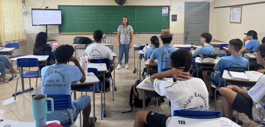 Fotografia de uma mulher em pé palestrando para jovens uniformizados em uma sala de aula com par...