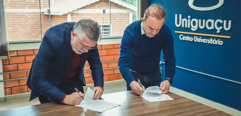 Uniguaçu adere ao Projeto Universidade Amiga da Justiça Eleitoral do Paraná Fotografia mostrando dois homens, um ao lado do outro, assinando documentos. Eles estão em uma s...