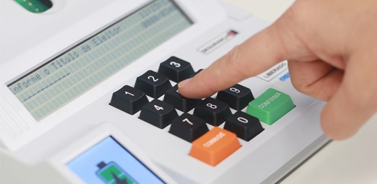 Foto de uma urna eletrônica, com a mão de uma pessoa clicando, com o dedo indicador, no botão ce...