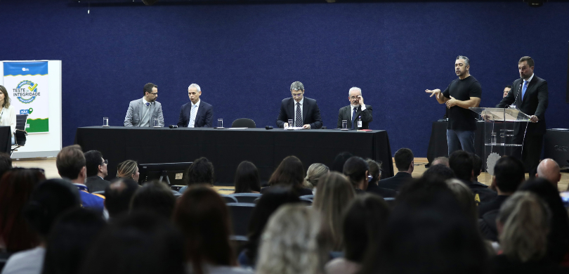Fotografia em ambiente interno na qual se observa o auditório lotado com diversas pessoas sentad...