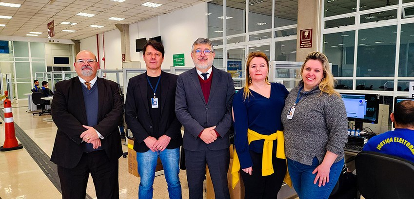 Urnas eletrônicas são preparadas para as eleições no Paraná Fotografia de cinco pessoas lado a lado. Os três primeiros são homens e usam terno. As duas últi...