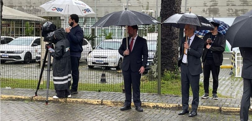 Fotografia em ambiente externo com piso molhado durante um dia de chuva. No centro, dois homens ...