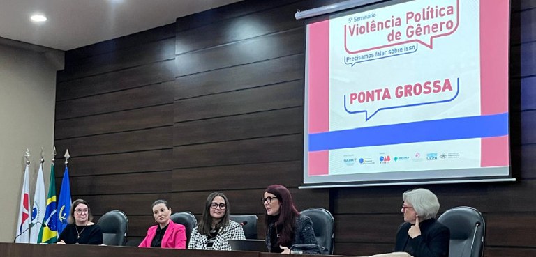 Fotografia de uma mesa de autoridades, composta por cinco mulheres, que estão sentadas em cadeir...