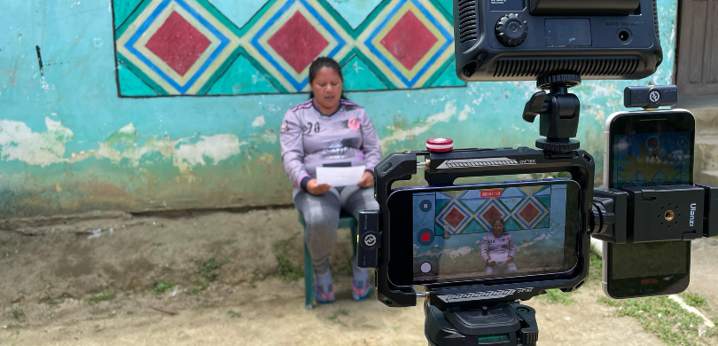 Vídeos e áudios no idioma Guarani Mbya levam informações sobre o processo eleitoral para pessoas indígenas Fotografia de uma mulher indígena sentada em uma cadeira verde, lendo um texto em uma folha de p...