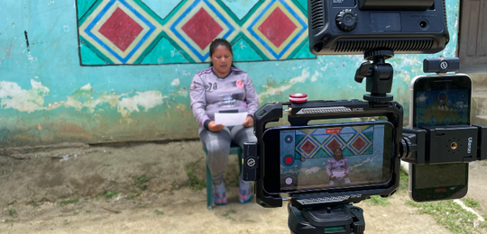 Fotografia de uma mulher indígena sentada em uma cadeira verde, lendo um texto em uma folha de p...