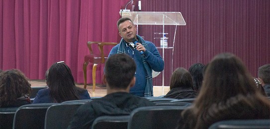 Fotografia de um homem falando em frente a um grupo de pessoas em um auditório. Ele usa uma blus...
