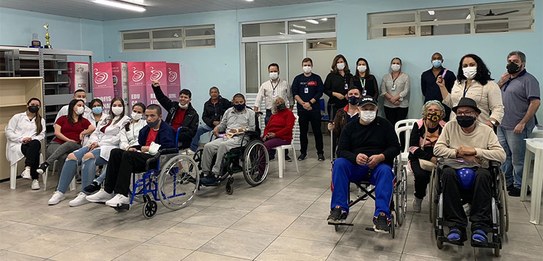 Fotografia de um grupo de pessoas em uma sala com paredes azuis. Algumas pessoas utilizam cadeir...