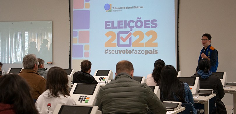 Fotografia de uma sala de aula, com pessoas sentadas com urnas eletrônicas à sua frente. Um home...