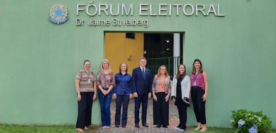 Fotografia na qual é possível ver dez mulheres e cinco homens posando sorrindo. Ao fundo, está a...