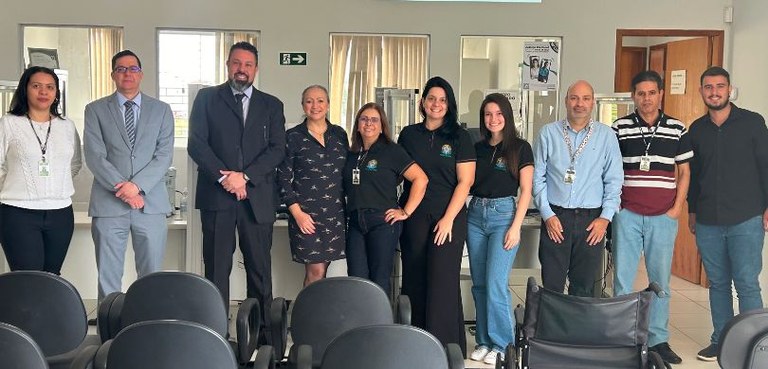 Fotografia de cinco homens e cinco mulheres sorrindo atrás de cadeiras com estofado preto enfile...