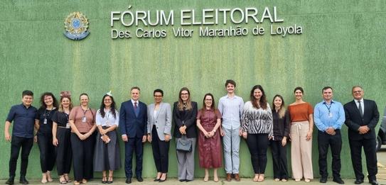 Fotografia na qual é possível ver dez mulheres e cinco homens posando sorrindo. Ao fundo, está a...