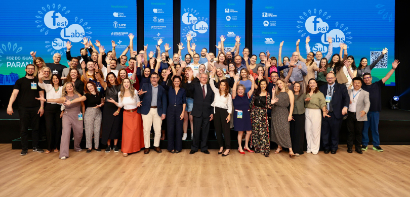 Fotografia de homens e mulheres posando sorrindo com os braços abertos. Algumas pessoas aparecem...
