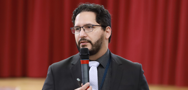 Fotografia de um homem de terno preto, camisa preta e gravata cinza. Ele segura um microfone com...